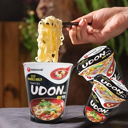 Cup of Nongshim Udon noodle soup with a hand lifting noodles, surrounded by additional Udon noodle cups.
