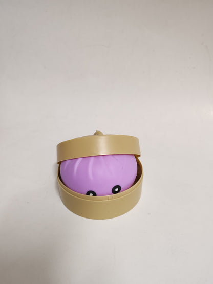 A small purple rounded toy or container with a beige lid, shown open on a plain light background. The front has two small black eyes.