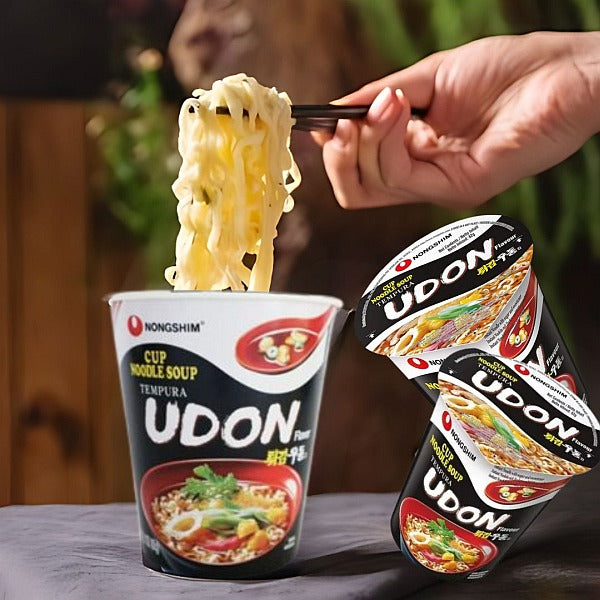 Cup of Nongshim Udon noodle soup with a hand lifting noodles, surrounded by additional Udon noodle cups.