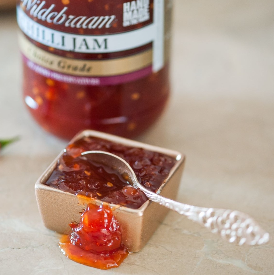 Jar of Wildbraam Chilli Jam with a spoonful of jam on a wooden surface