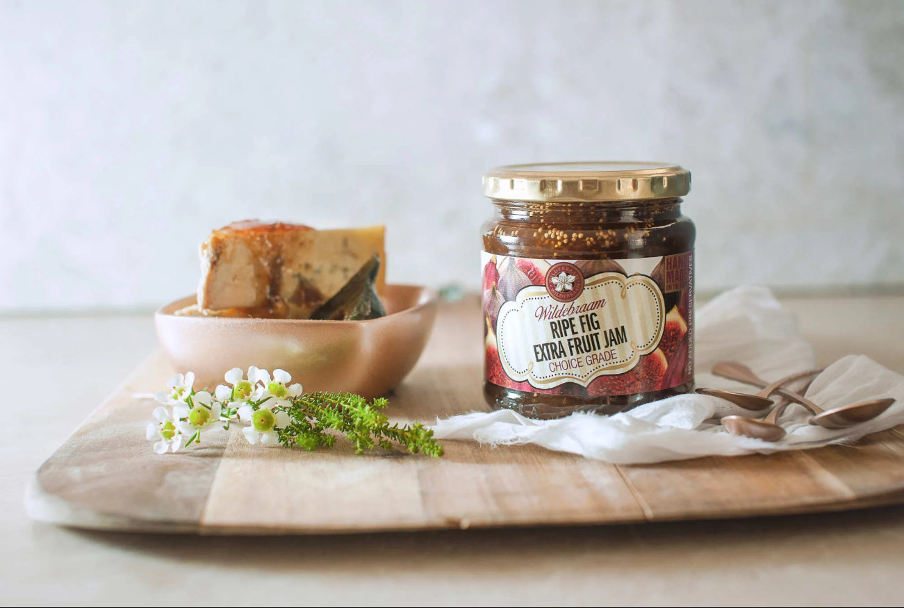 Jar of Nelly's Fig Extra Fruit Jam on a wooden board with a bowl of figs and flowers.