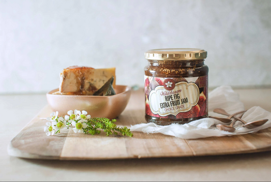 Jar of Nelly's Fig Extra Fruit Jam on a wooden board with a bowl of figs and flowers.