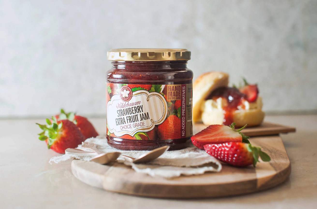 Jar of strawberry fruit jam on a wooden board with strawberries and bread in the background.